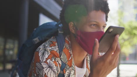 African-american-man-in-city,-wearing-face-mask-sitting-using-smartphone-in-street