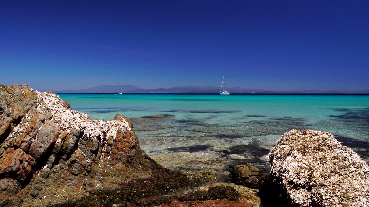 la cámara se acerca entre dos rocas en una hermosa playa con un velero en el fondo