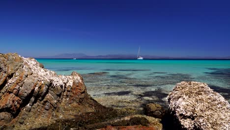 La-Cámara-Se-Acerca-Entre-Dos-Rocas-En-Una-Hermosa-Playa-Con-Un-Velero-En-El-Fondo