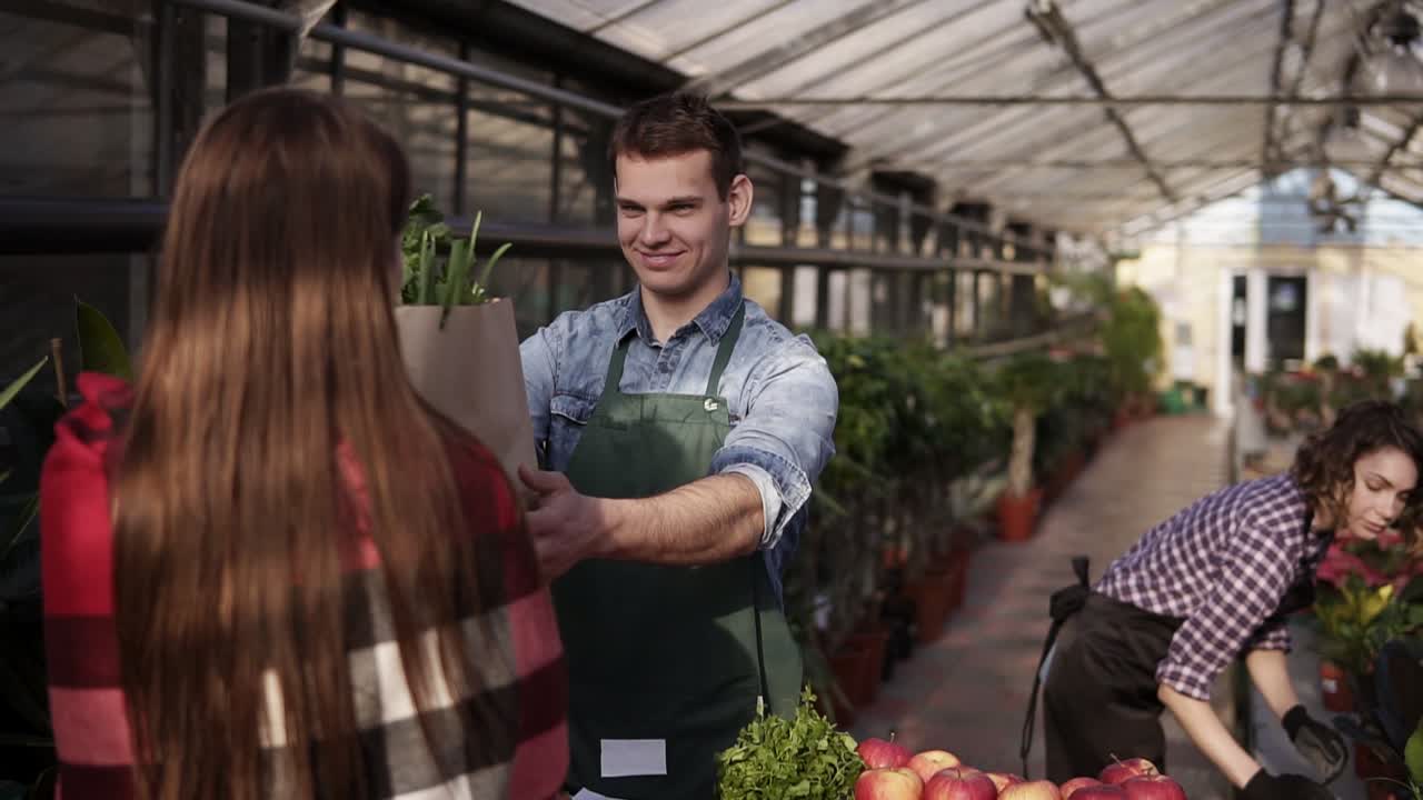 Portrait of european salesman wearing green apron is giving organic food in brown paper bag to female customer in greenhouse. Cheerful, smillling man selling organic food from the table while his wife harvesting on the background