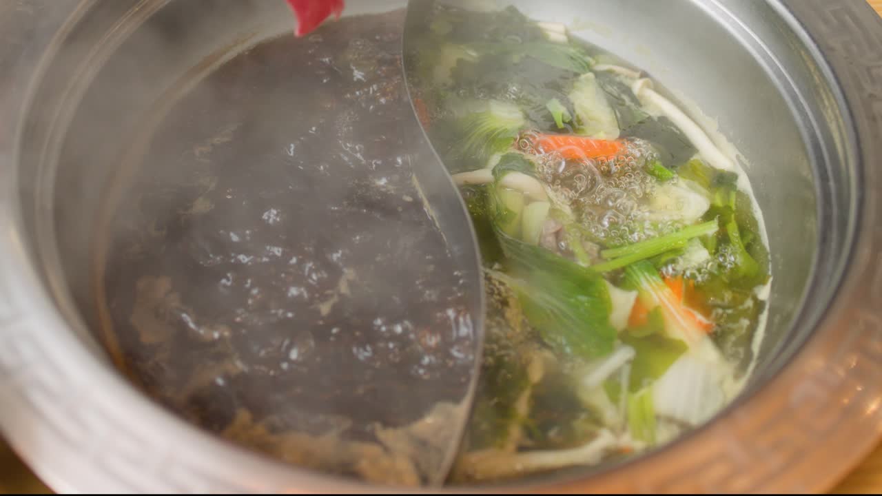 Hand dips raw beef into steaming dual broth hotpot, vegetables boiling, overhead view, warm lighting