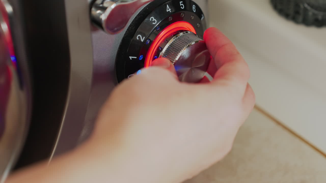 Close up of hand adjusting silver control knob of electric stand mixer with red and blue light indicators in cozy kitchen, showcasing operation of appliance in cooking or baking environment