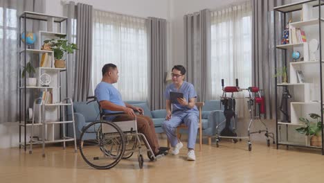 vista lateral completa del cuerpo de un enfermero masculino asiático y un paciente en silla de ruedas hablando sobre los resultados de las pruebas médicas en una tableta después de la fisioterapia en casa