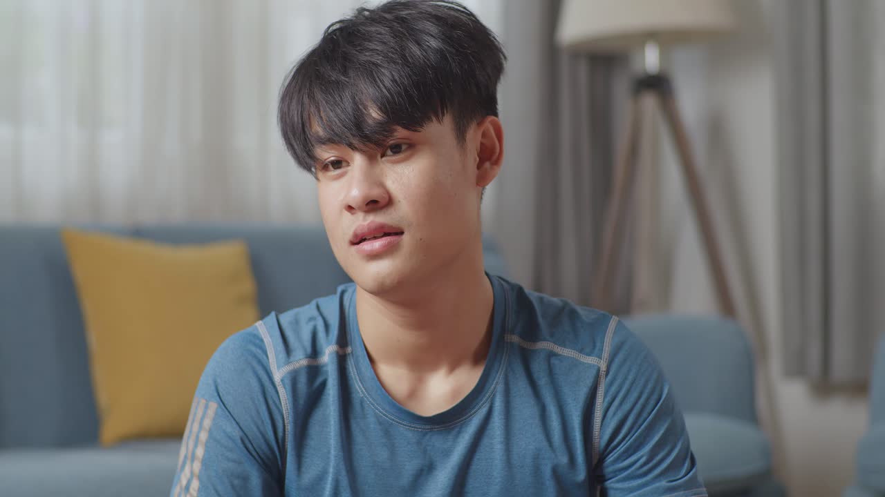 운동복을 입은 아시아 남자가 운동을 마친 후 집에서 미니멀한 인테리어를 가진 넓고 은 거실에서 포즈를 취하고 있습니다.