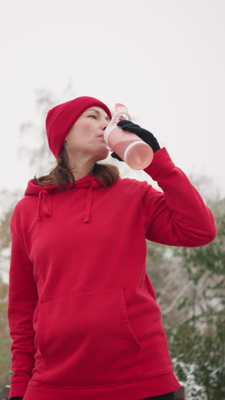 woman drinks water from pink bottle outdoors, lowering it while gazing into distance, serene winter setting with misty atmosphere, snowy trees, vintage streetlamp in background, peaceful moment