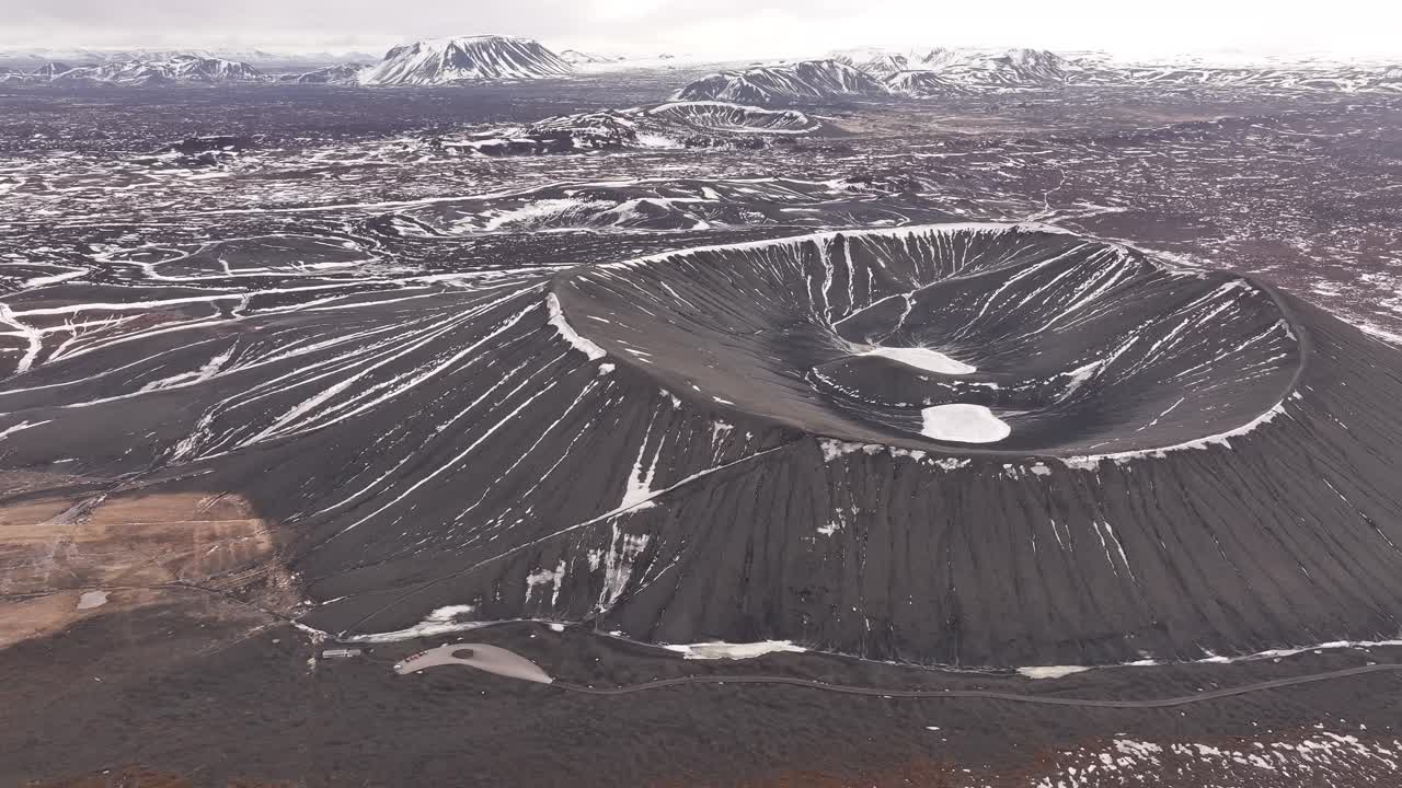 Majestic aerial view of Hverfjall crater near Lake Mývatn, Iceland. A stark volcanic cone dusted with snow stands amid rugged lava fields in a dramatic winter landscape.