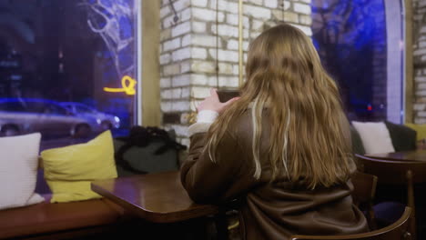 close up back view of student in brown shearling jacket sitting at cozy cafe sipping coffee, with people walking outside window. soft lighting adds calm and peaceful ambiance to scene