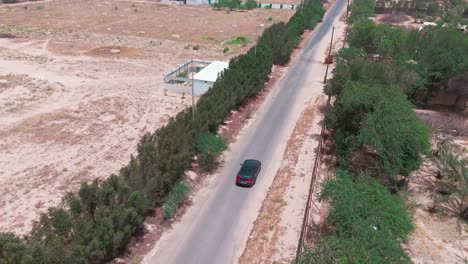 4k aerial footage of a black car cruising down an empty road surrounded by desert terrain, trees, and farm plots. ideal for travel, transport, isolation, or cinematic b-roll.
