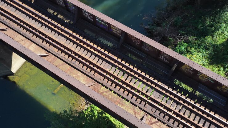 Train crossing railroad bridge with rusted metal supports and wooden planks, drone top down tracking over Old River in Benton Arkansas