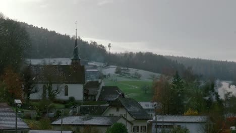 snow is falling over the rooftops of cavergno village while the sun is setting behind the dark forest in the background