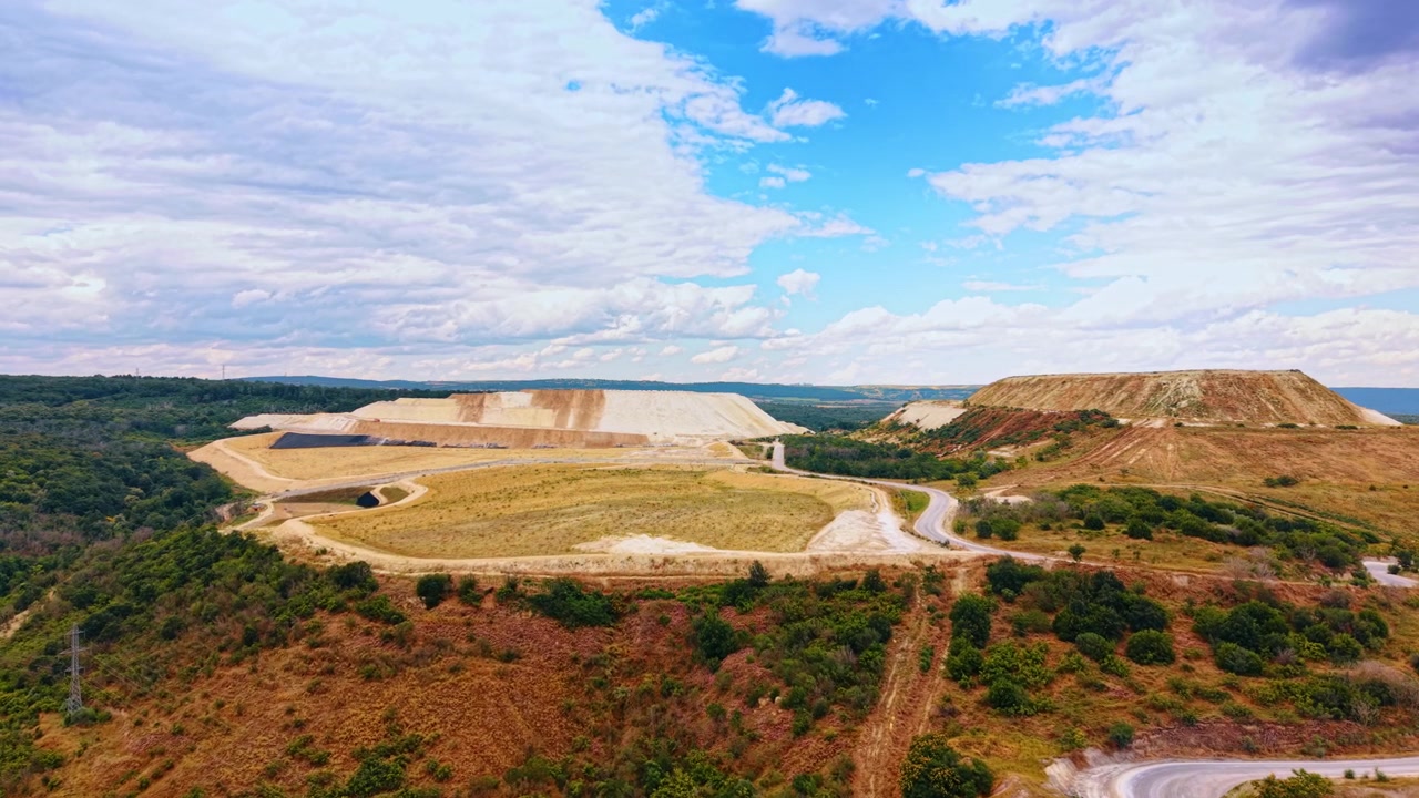 Vista aérea de uma paisagem de pedreira a céu aberto em socalcos. Uma estrada sinuosa serpenteia por montículos de terra bege rodeados por árvores verdes sob um céu azul nublado.