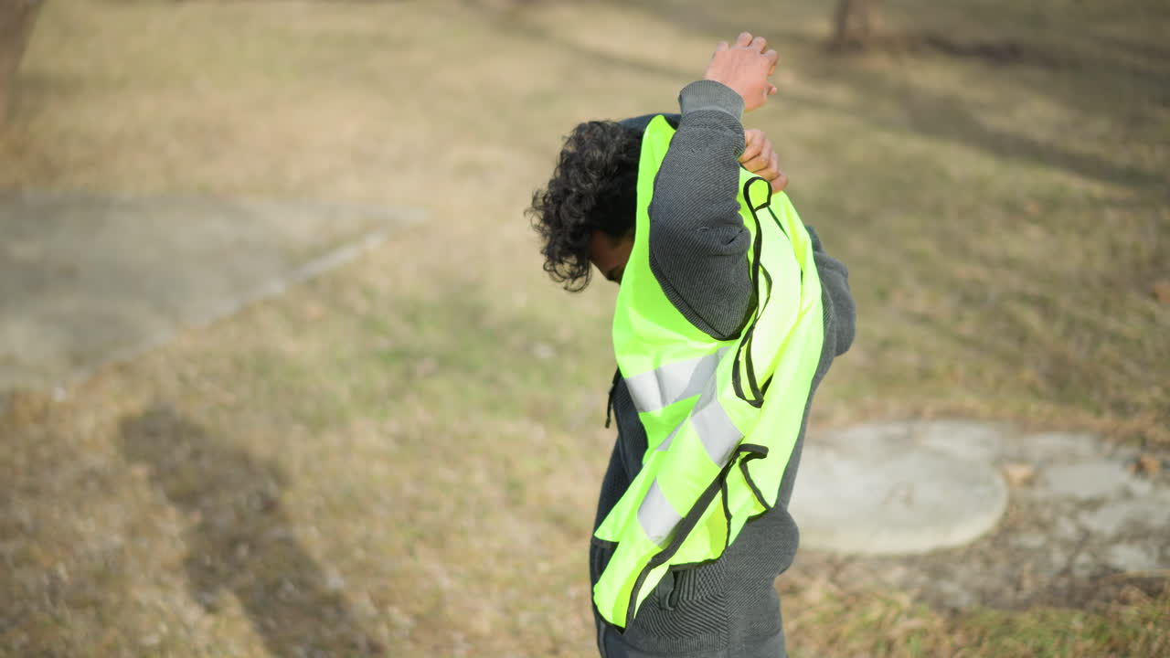 Man outdoors putting on bright neon green reflective safety vest, raising arm to slip through opening, preparing for outdoor work or volunteer activity in park with bare trees during early spring