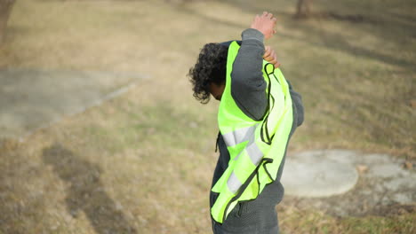 man outdoors putting on bright neon green reflective safety vest, raising arm to slip through opening, preparing for outdoor work or volunteer activity in park with bare trees during early spring