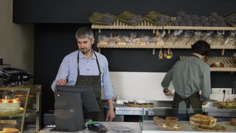 Handsome-father-in-apron-passes-a-tray-of-pastries-to-his-young-son-and-cashier-go-to-the-checkout.-Indoor,-close-up,-slow