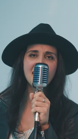 cantante femenina apasionada con sombrero negro y chaqueta de cuero actuando intensamente en un micrófono vintage, con un fondo blanco suave