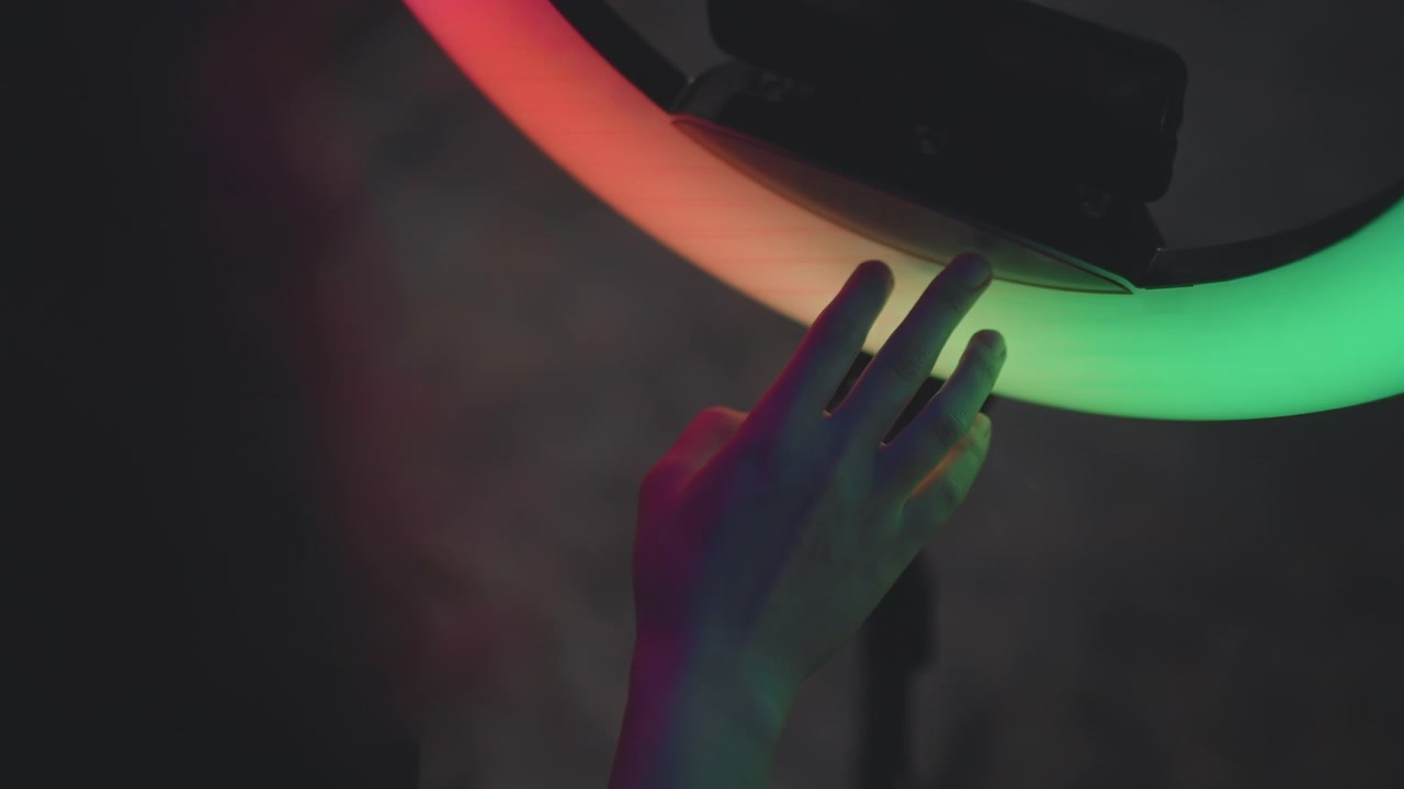 Nærbillede af hånd, der rører ved en delvis cirkulær ringlampe, mens farver skifter gennem orange, grønne, blå, lilla og hvide segmenter, med blød glød, let berøring og stemningsfuld studieatmosfære