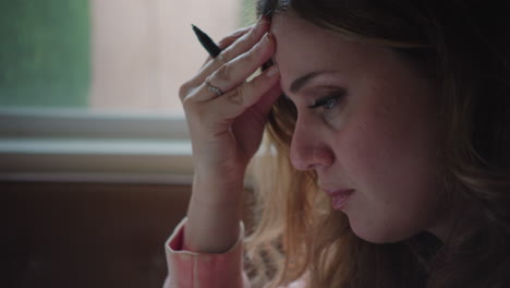 Handheld-Shot-of-Stressed-Out-Woman-Holding-Pen-at-Table,-Contemplating-and-Pensive-Beside-Window-in-the-Daytime