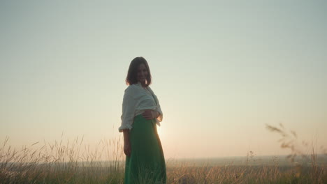 young adult stands in tall grass during golden hour, gracefully turning with gentle smile and placing hand on stomach while soft evening light casts glow across peaceful natural landscape