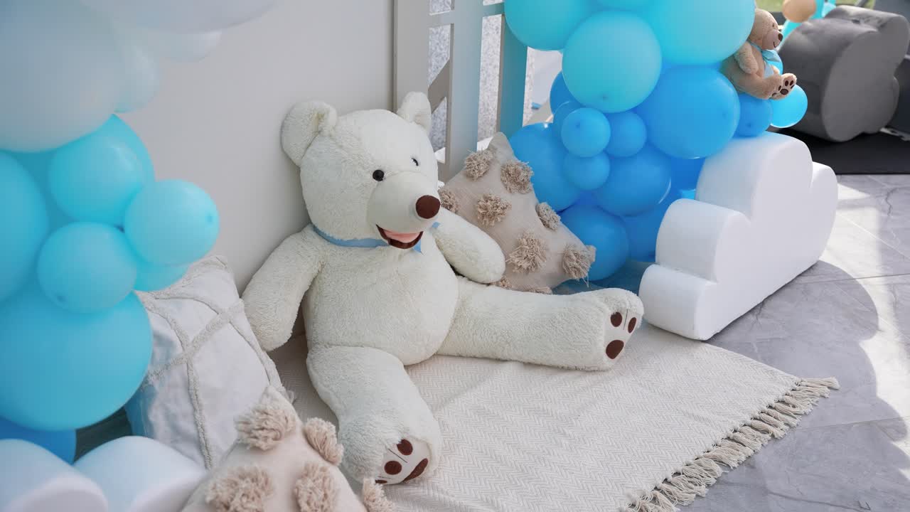 Adorable white teddy bear seated among blue balloons and pillows, creating a cozy and playful setup for a baby-themed celebration or christening event