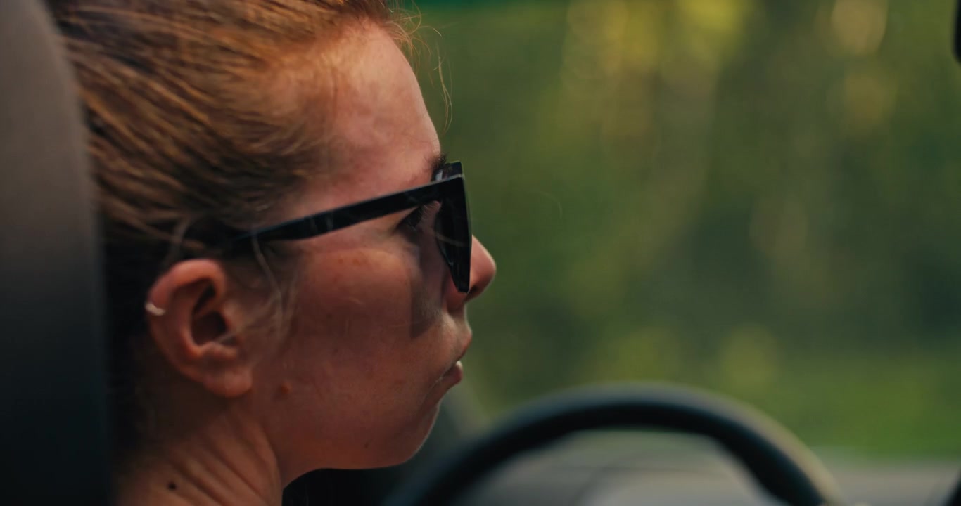 Over the shoulder view of a woman driving through a forest wearing sunglasses. Soft woodland background and warm natural light create a relaxed summer road trip atmosphere.