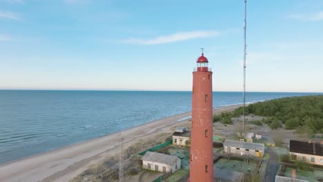 luftaufnahmen des rot gefärbten leuchtturms von akmenrags, der ostseeküste, lettlands, des weißen sandstrandes, der ruhigen see, des sonnigen tages mit wolken, einer weiten drohnenaufnahme, die sich vorwärts bewegt