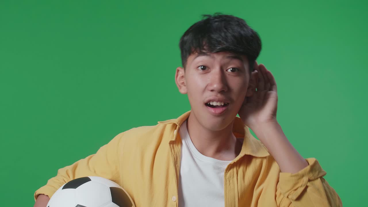 초록색 스크린 배경에서 축구를 응원하는 동안 듣기 위해 공을 고 있는 아시아 소년의 근접 사진