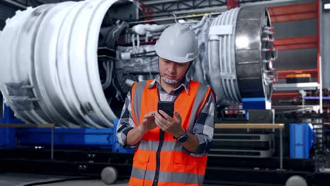 asian male engineer with safety helmet using smartphone and looking around while standing with airplane engine maintenance conducted, analytics checking the turbine