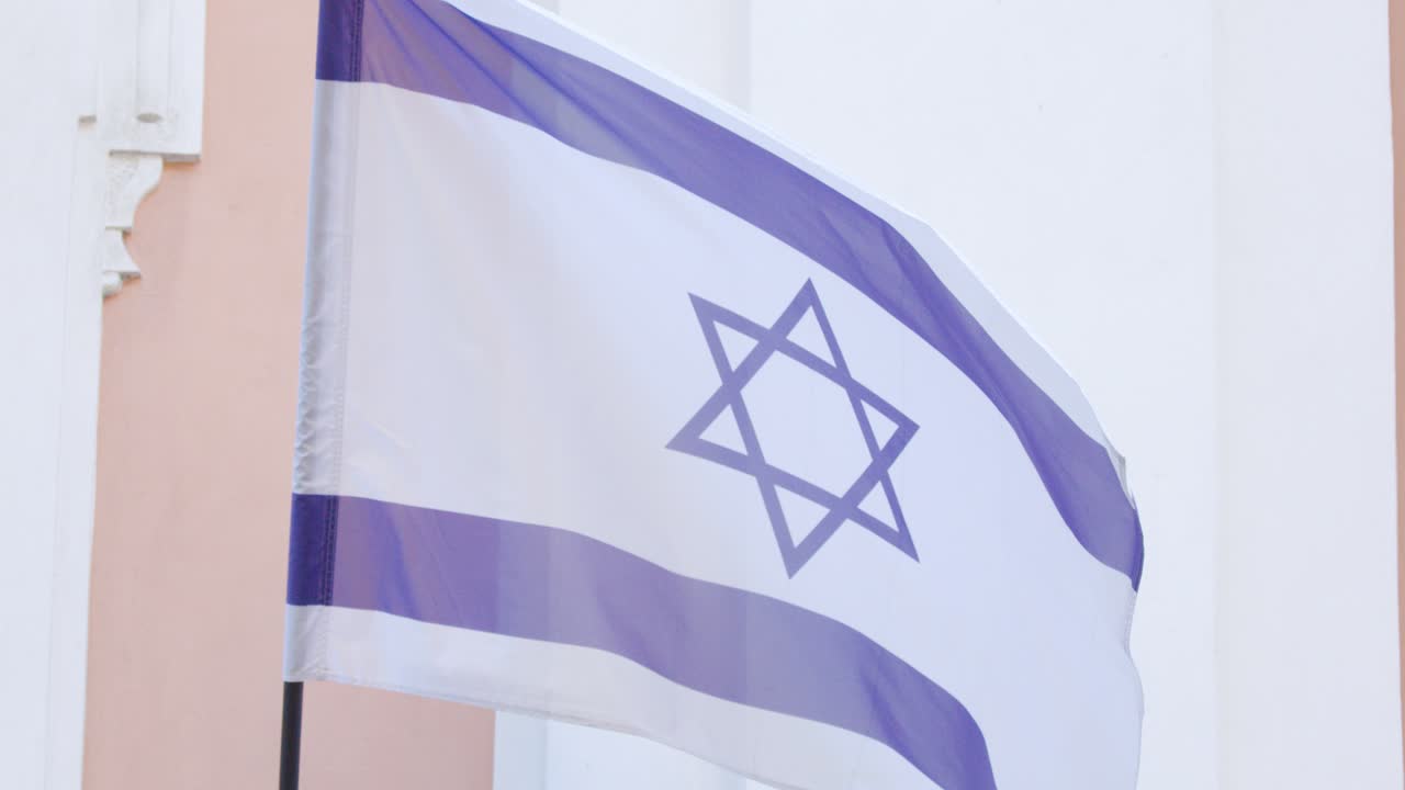 Bandiera israeliana sventola sull'asta, luce diurna soffusa, telecamera statica, sfondo di un edificio pallido, atmosfera calma.