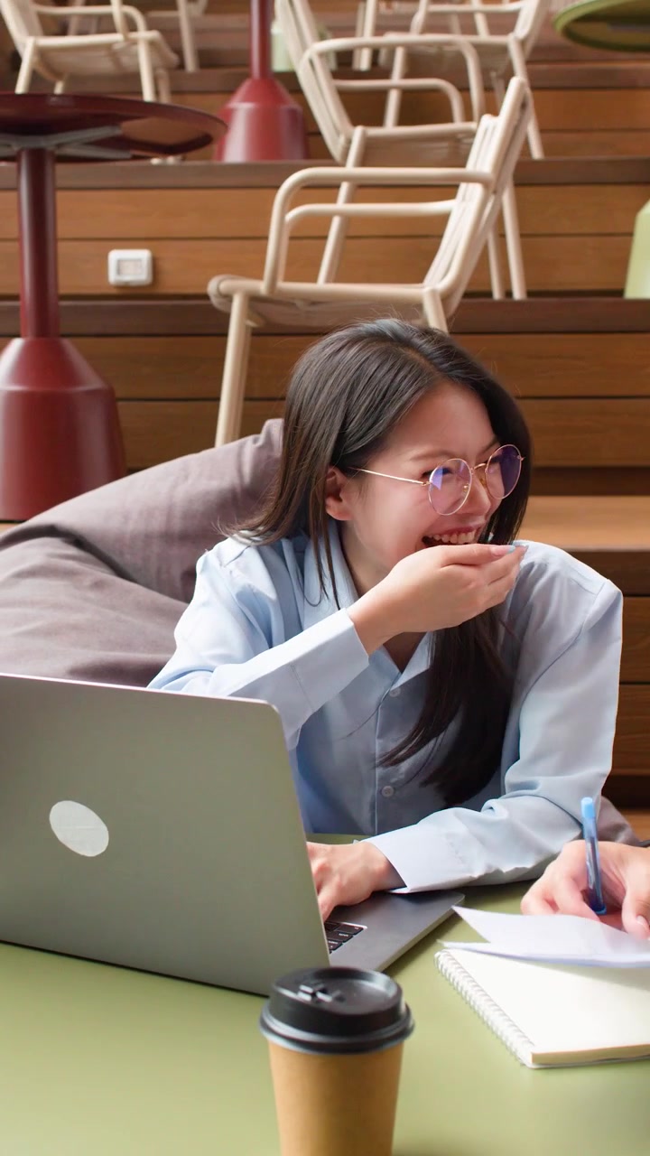 Ung asiatisk forretningskvinne med briller diskuterer arbeid med kollega mens hun bruker laptop i en moderne lounge