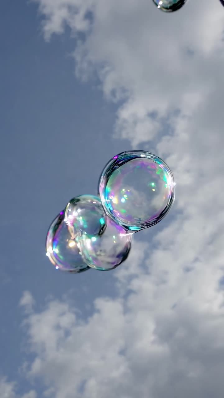 A low-angle video captures iridescent soap bubbles floating against a cloudy sky