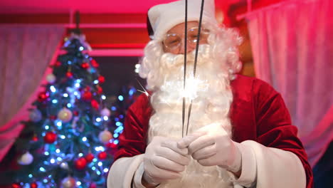 santa indoors on new year tree background. portrait of a real santa claus with white beard holding sparkling lights and looking at camera. happy new year.