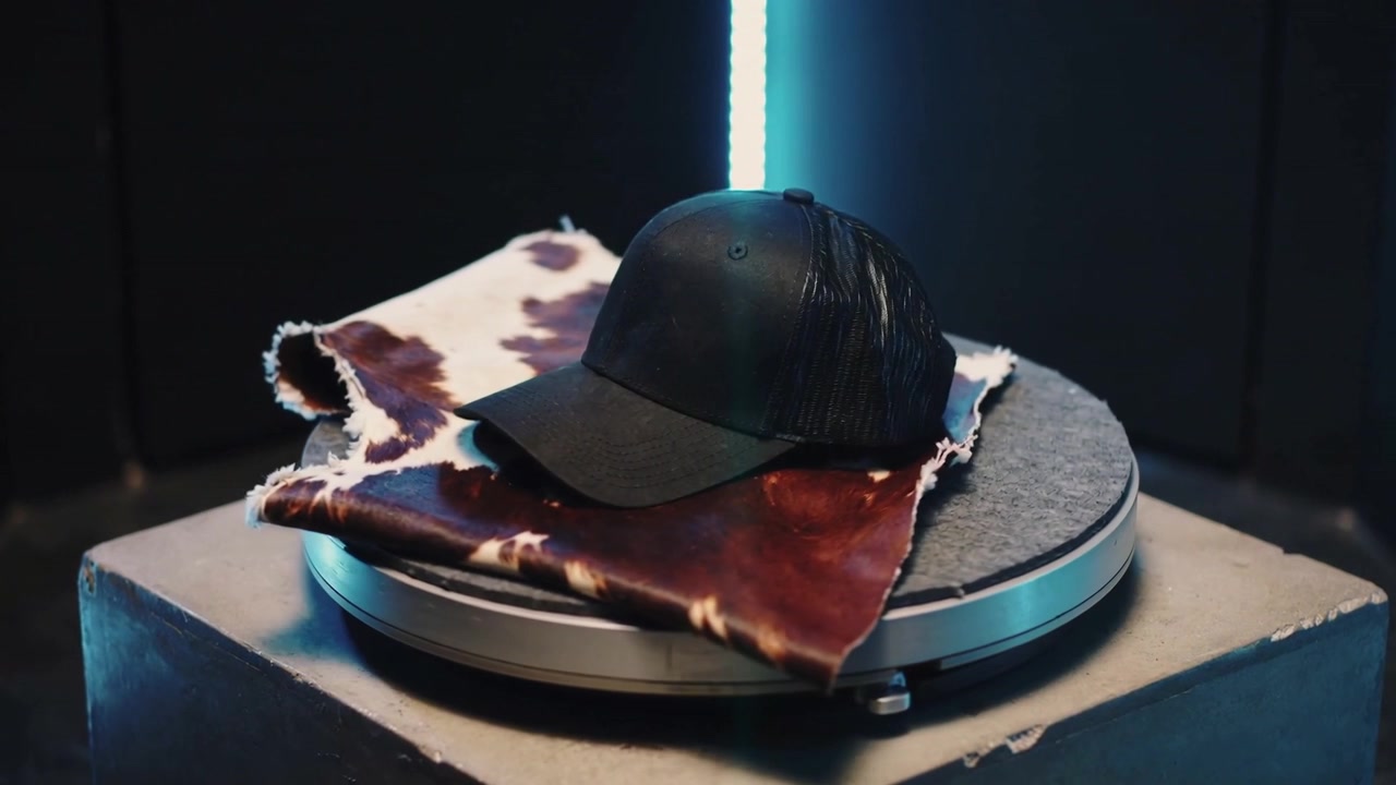 Piattaforma girevole che trasporta un cappellino nero, rivelando angolazioni per mostrare il prodotto in studio, con sfondo in pelle di mucca e LED blu.