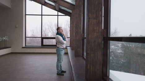 man looks at winter landscape out of panoramic window. thoughtful mature gentleman stands alone by glass wall in residential house. calm solitude