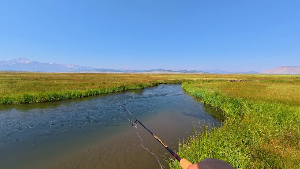First-person shot of an angler casting a spinning rod into a clear, meandering river bordered by lush green grasses. Wide open Owens Valley landscape with the Sierra Nevada mountains on the horizon