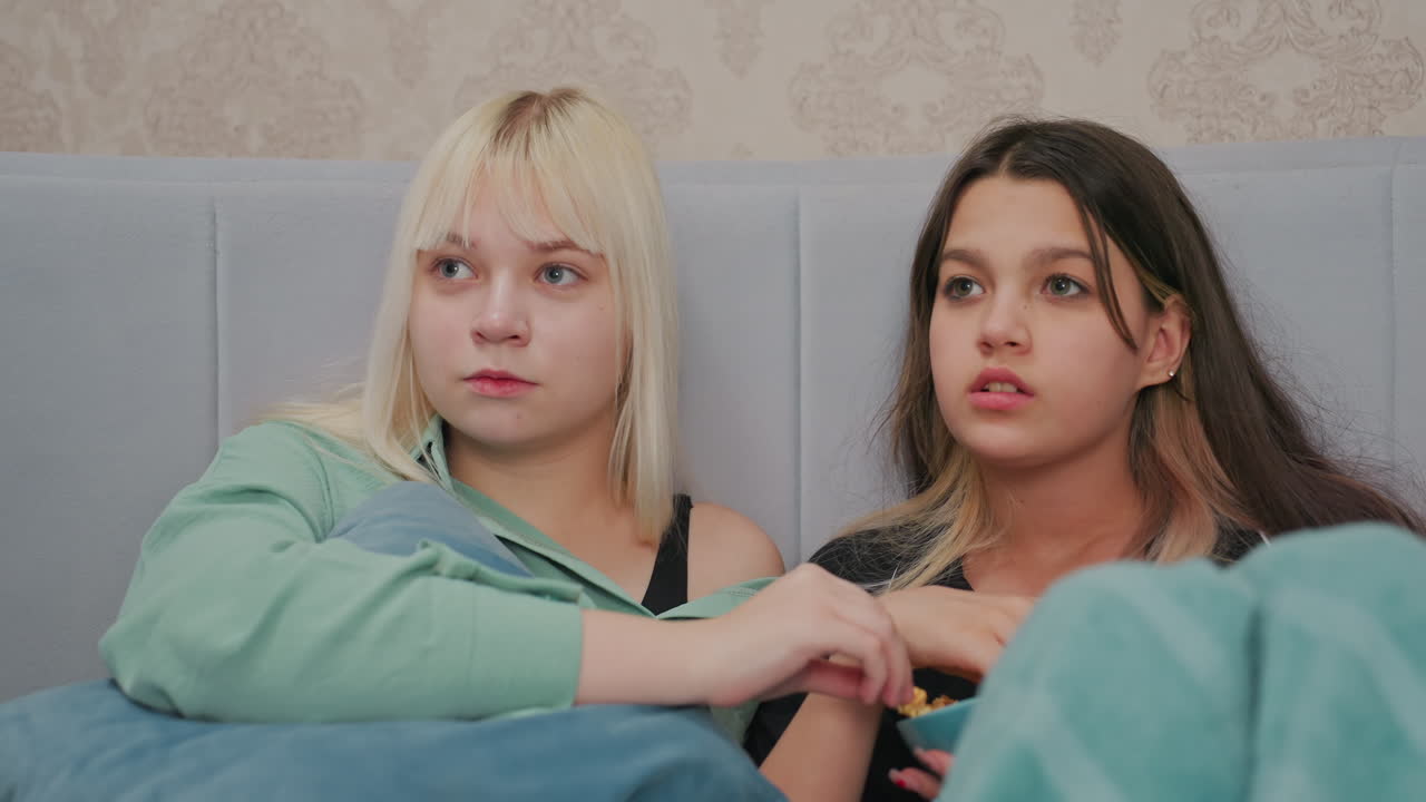 two young girls sitting on bed watching movie with focused expressions while sharing bowl of popcorn, one girl holding pillow, other eating popcorn, cozy atmosphere with teal blanket