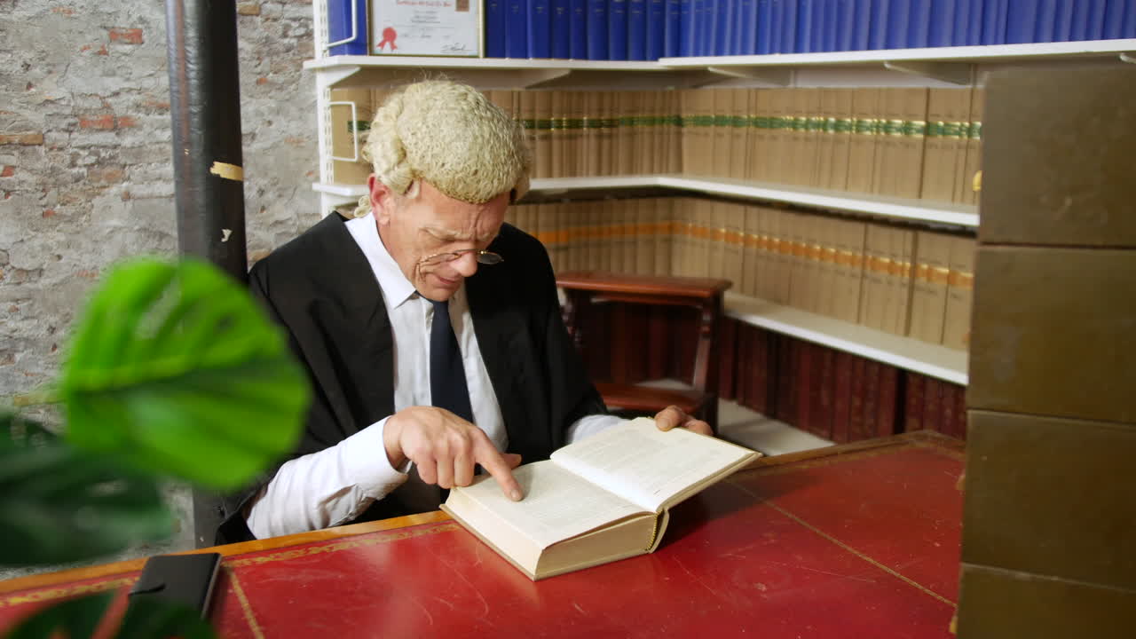 Dolly shot of judge or barrister reading a law book in the judge's chambers library. The man is wearing a wig and gown with glasses