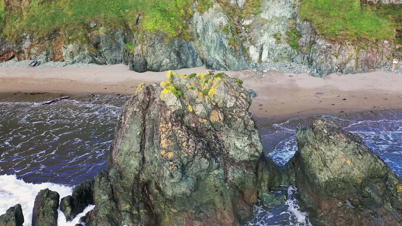 Orbitalluftfoto af en farverig klippe langs Irlands sydkyst med bølger, der bryder omkring den.