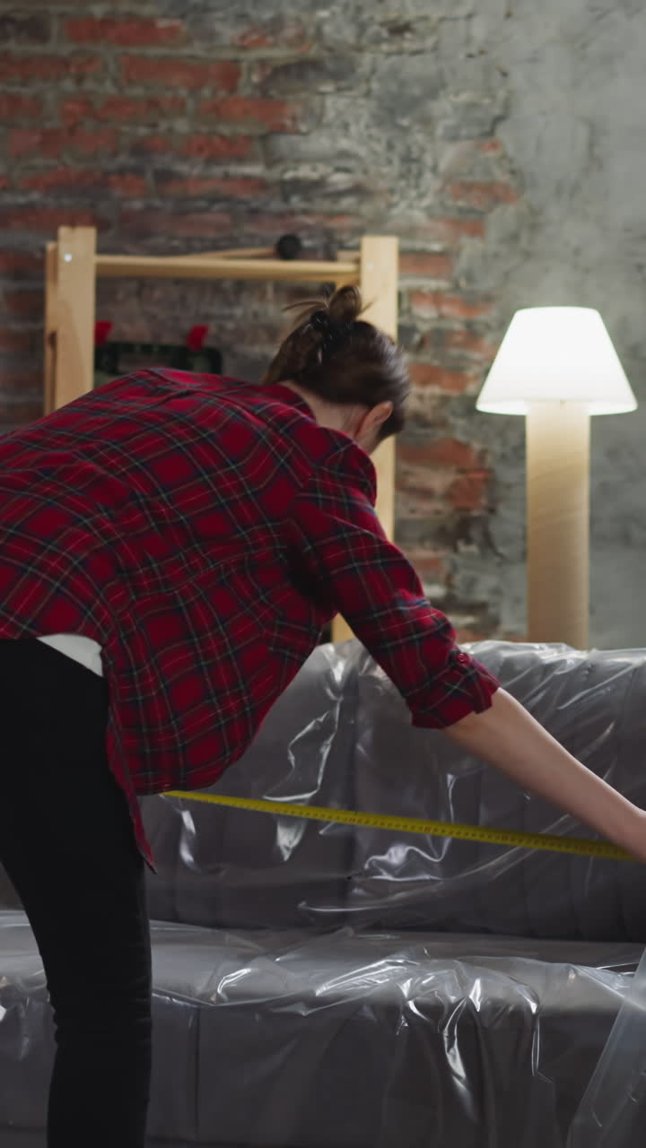Woman measures sofa length covered with foil using tape in living room backside view. Lady works in room prepared for renovation. Home reconstruction
