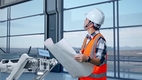 vista lateral de un ingeniero masculino asiático con casco de seguridad mirando el plano en sus manos y mirando a su alrededor mientras está de pie en una fábrica de alta tecnología, analizando el concepto de máquina robótica