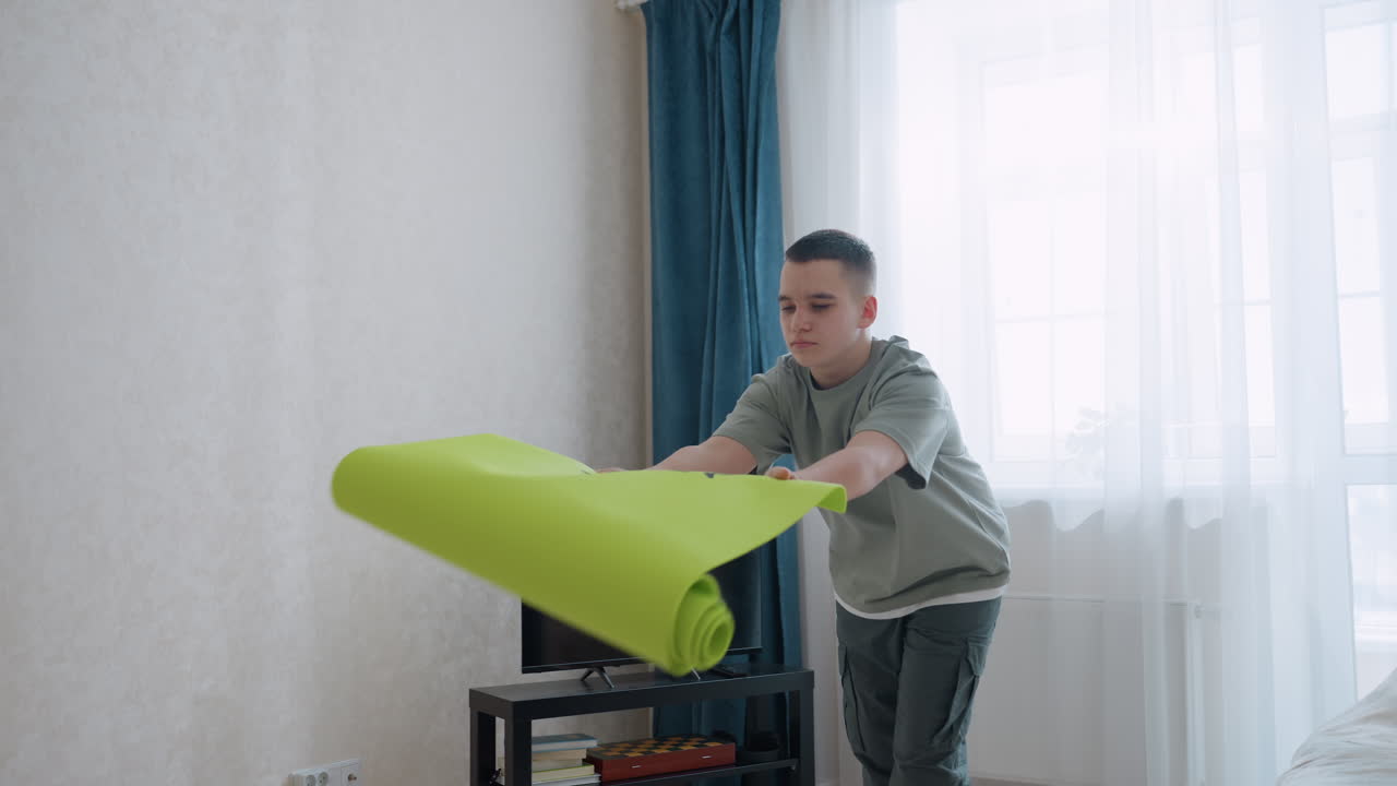 Boy dressed in casual light green outfit carefully unrolls bright green fitness mat on wooden floor inside sunlit room with curtains and TV stand