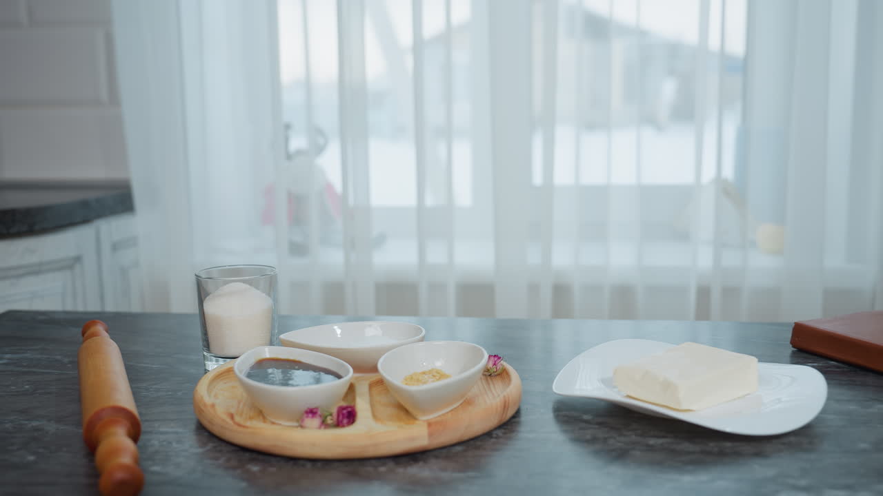 주방 필수품, 설탕, 버터, 갈색 책과 함께 나무 트레이에 있는 접시에 담긴, 색 투명한 커튼은 자연빛을 허용하며 눈으로 인 건물을 드러니다.