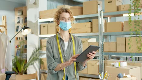 Young-caucasian-designer-woman-in-facial-mask,-typing-on-tablet-and-looking-at-camera-at-office-in-clothing-shop