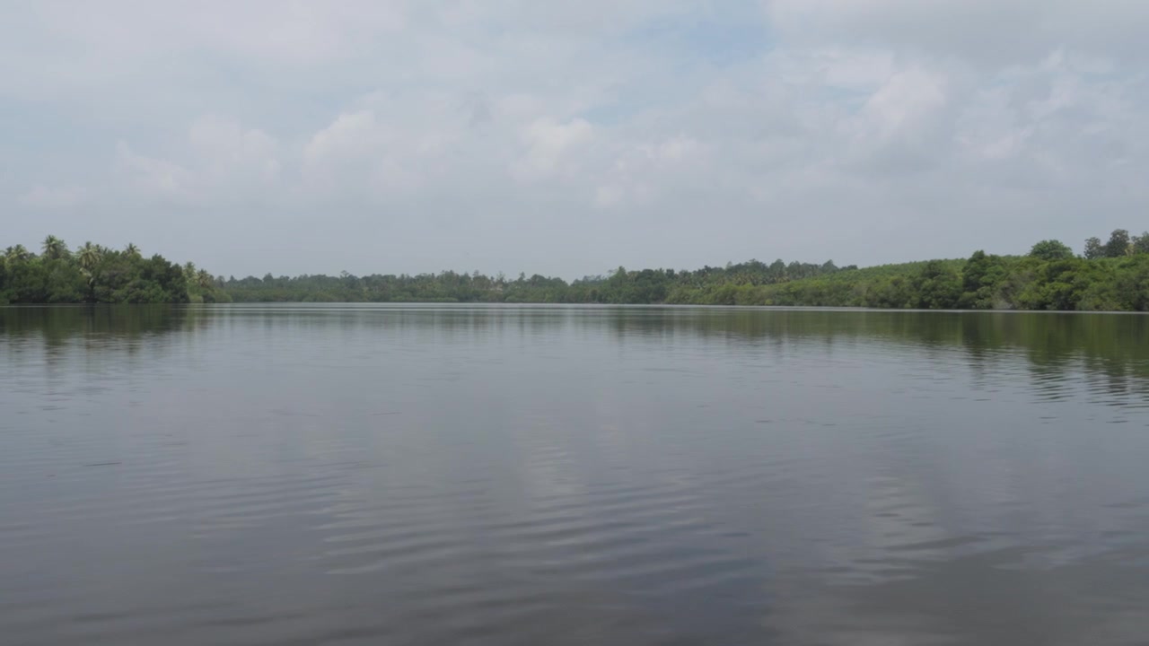 Wide view of the calm Hikkaduwa River with soft ripples, green tropical banks, distant palms and cloudy daylight atmosphere in Sri Lanka.