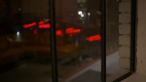 Looking-out-the-window-of-industrial-building-with-red-and-purple-glowing-city-lights-out-of-focus-in-background,-and-reflection-of-high-end-yellow-couch-in-glass