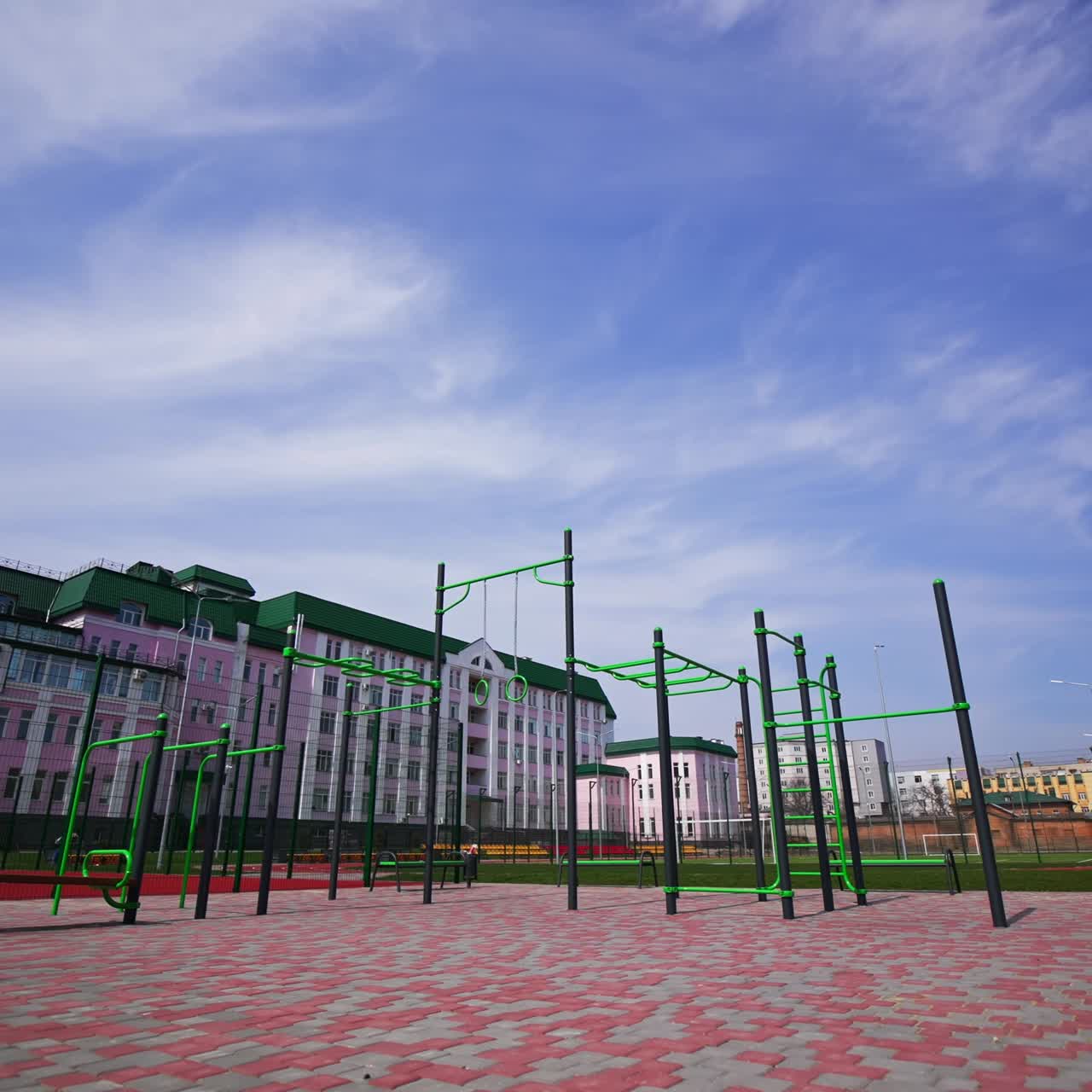 Approaching the set of sport horizontal bars at the outdoor sport ground with paved floor. Running tracks, football field and long pink building at backdrop