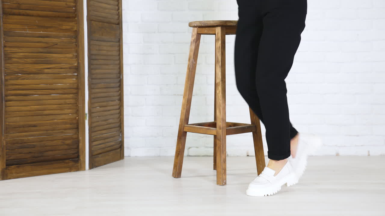 Model in black jeans demonstrating white massive shoes. Lady turns around to show the fashionable footwear. Wooden tabouret at backdrop.