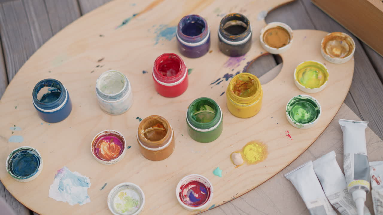 palette in legno dai colori vivaci con barattoli di vernice disposti su un tavolo in legno all'aperto, tubetti di vernice sparsi e pigmenti acrilici colorati, primo piano di un set creativo per pittura e schizzi su strada, macchie materiche