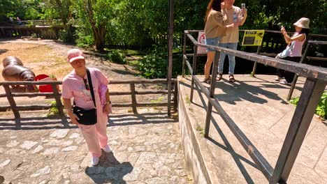 a hippopotamus approaches a red bucket in a zoo enclosure under bright sunlight, observed by visitors