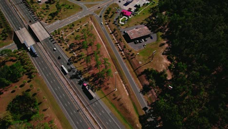 Aerial-view-of-trucks-on-highway,-near-logistics-hub-with-warehouses-and-green-landscape