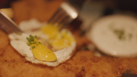 close-up of a fork and knife slicing a fried egg atop golden chicken schnitzel, with creamy sauce on the side under warm, soft lighting